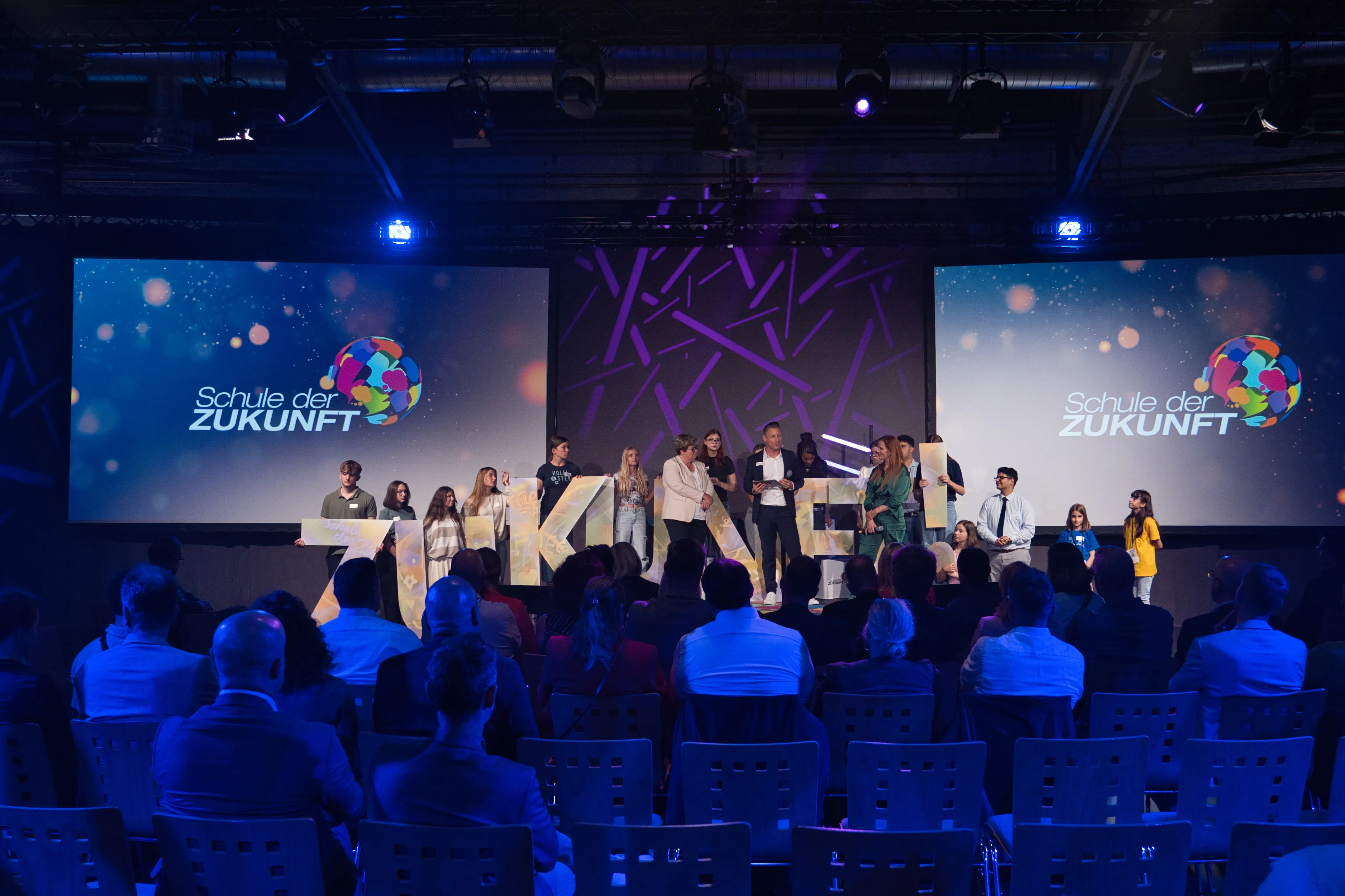 Eine Gruppe von Schüler:innen steht auf einer Bühne und hält Buchstaben in der Hand, die das Wort „ZUKUNFT!“ bilden